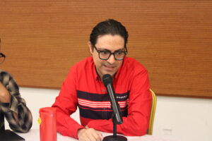 Hombre con camisa roja sentado, exponiendo algún tema.
